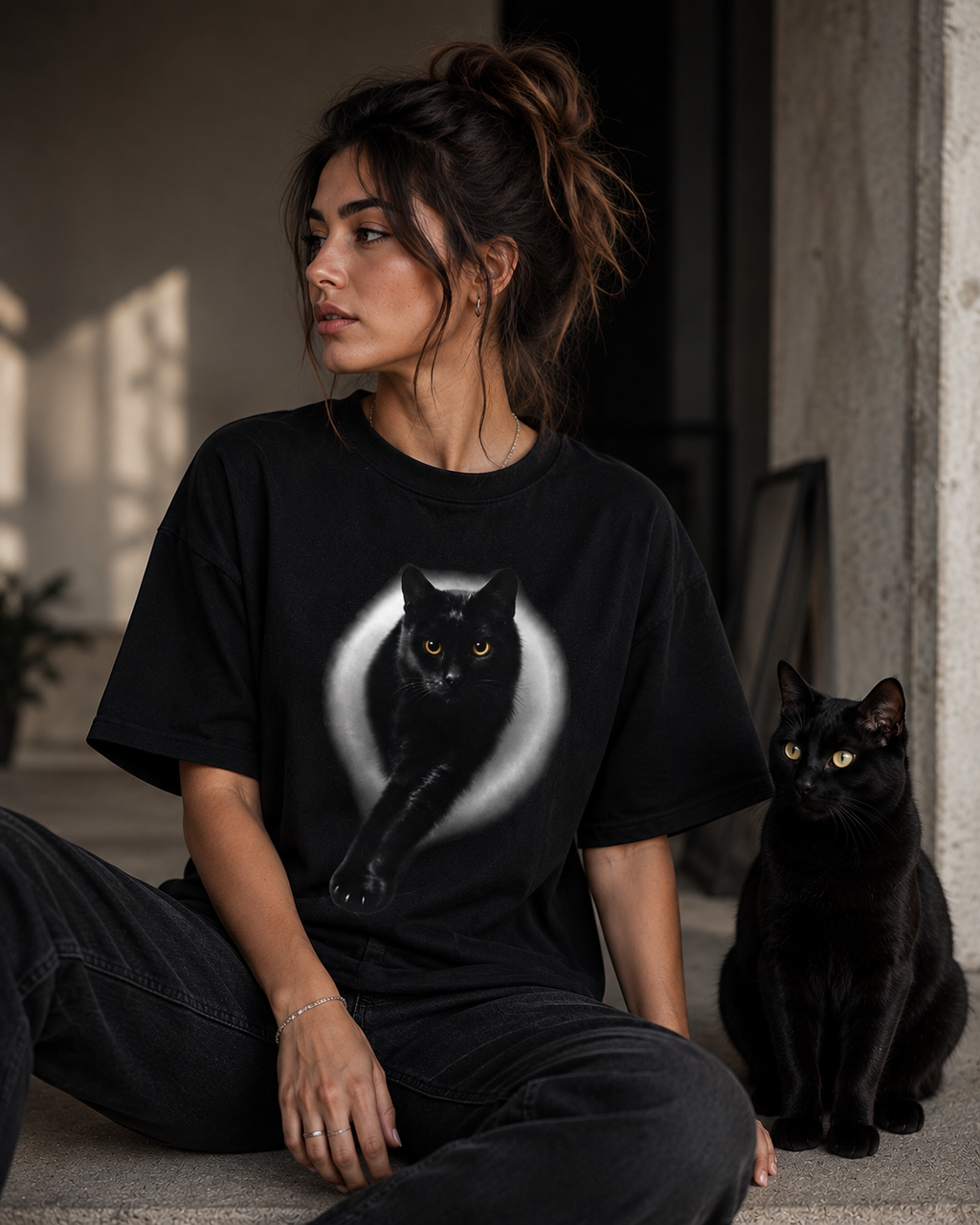 Woman wearing black cat graphic t-shirt sitting beside a real black cat, realistic cat print on premium oversized black cotton tee for cat lovers