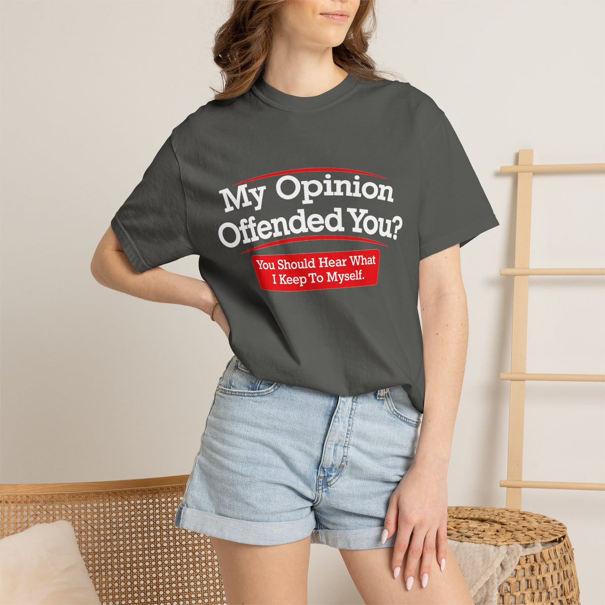 A woman wearing a dark charcoal gray graphic t-shirt with the text 'My Opinion Offended You? You Should Hear What I Keep To Myself!' in white and red, paired with light-wash denim shorts in a neutral indoor setting.